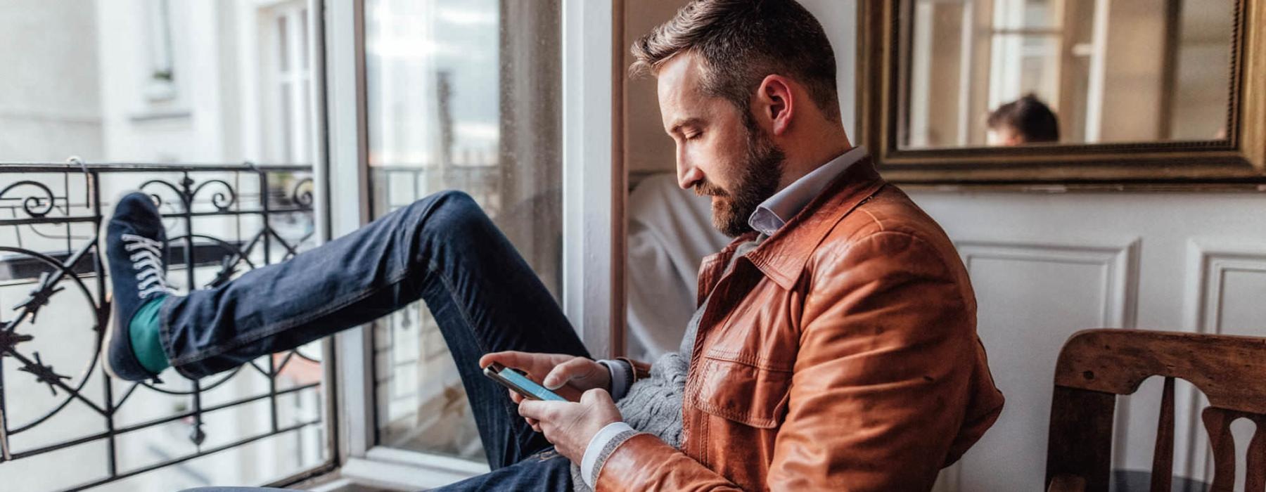 a man sits on a chair and rests his foot on a balcony railing as he looks at his phone in his room